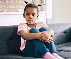 child in pink tshirt, blue jeans and pink socks sitting on grey sofa looking at camera