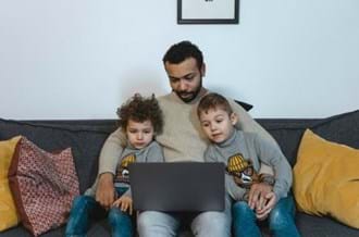 2 children and an adult sitting on a sofa at home looking at a laptop.