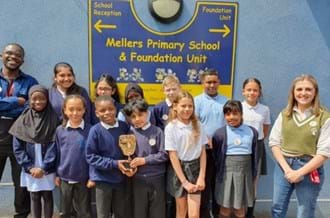 A group of Mellers Primary School students smiling and standing with BAFTA Roadshow presenters De-Graft Mensah (left) and Kia Pegg (right) in front of the school sign. 