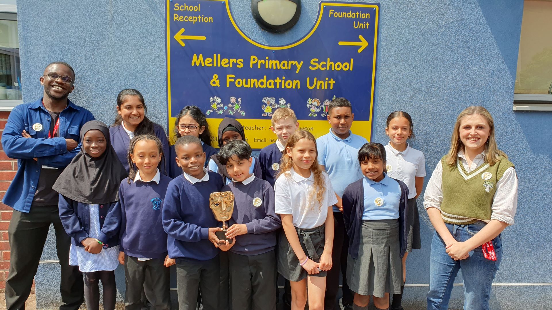 A group of Mellers Primary School students smiling and standing with BAFTA Roadshow presenters De-Graft Mensah (left) and Kia Pegg (right) in front of the school sign.