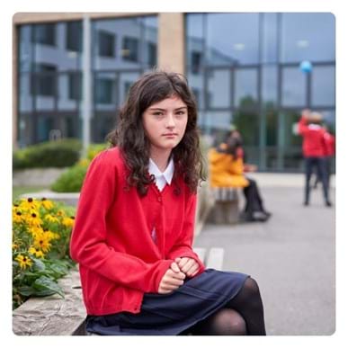 A young girl in red school uniform sitting on a bench outside her school, looking into the camera.