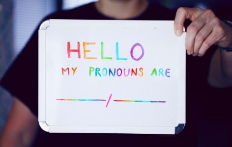 A person holding a whiteboard that says HELLO, MY PRONOUNS ARE in rainbow colours