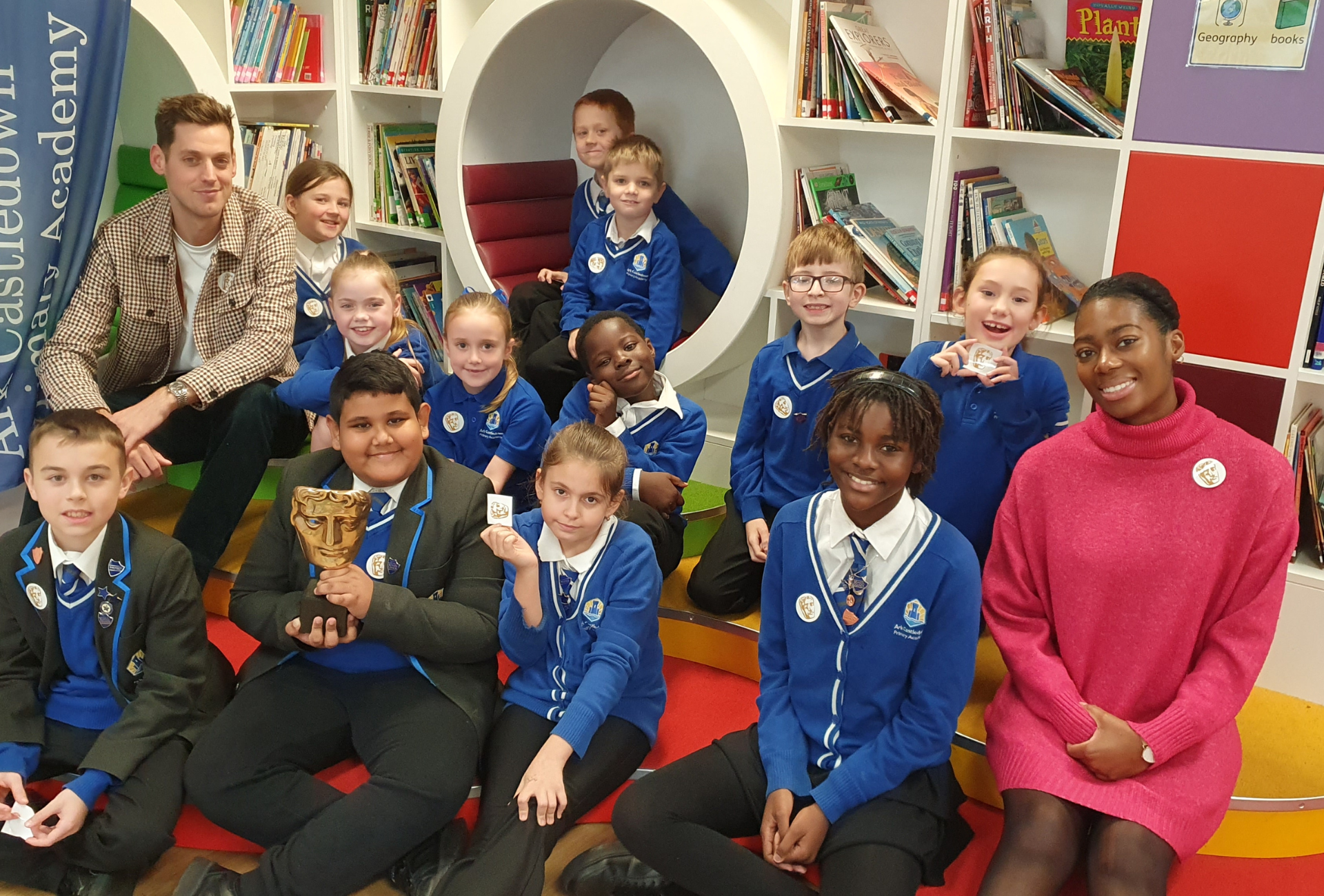 BAFTA Roadshow presenters Sam Homewood (left) and Shanequa Paris (right) in a classroom with a group of smiling children from Ark Castledown Primary Academy in Hastings.