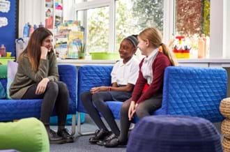 A qualified Place2Be counsellor talking to two school pupils in a classroom environment. They are all sitting on blue sofas and smiling.