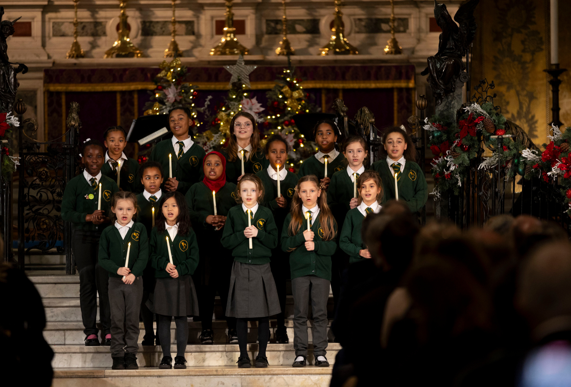 The Goldfinch Primary School choir performing at Holy Trinity Church in Sloane Square. Fifteen children dressed in school uniform are holding candles while singing.