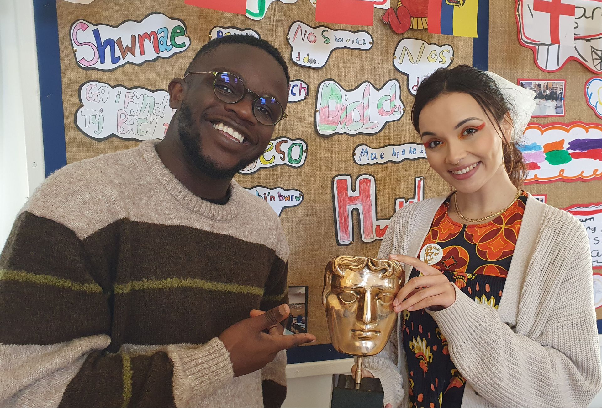 De-Graft Mensah and Mirain Iwerydd standing in a classroom smiling while holding a BAFTA trophy.