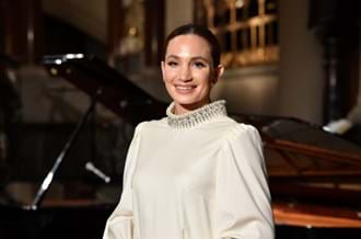 Award winning mezzo-soprano, composer and long-time Place2Be supporter Laura Wright smiling as she stands in front of a piano. Her hair is pulled back and she is wearing a cream shirt.  